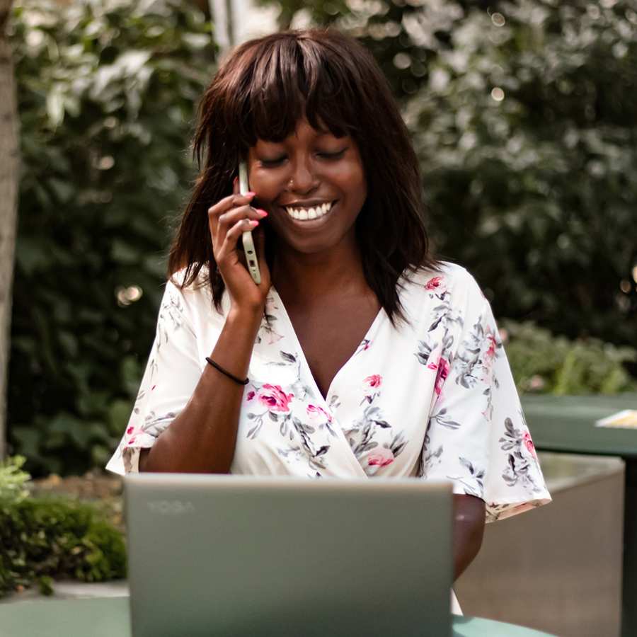 Ina met telefoon en laptop, glimlachend, met wit bloemenjurkje